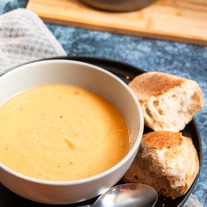 a bowl of parsnip soup on a black plate with crusty bread and a silver spoon. A pot of soup with a wooden spoon on a wooden board in the background,