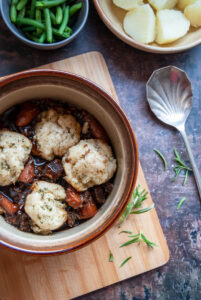 a brown ceramic dish of beef stew with carrots and herb dumplings, a bowl of green beans and potatoes and a silver spoon.