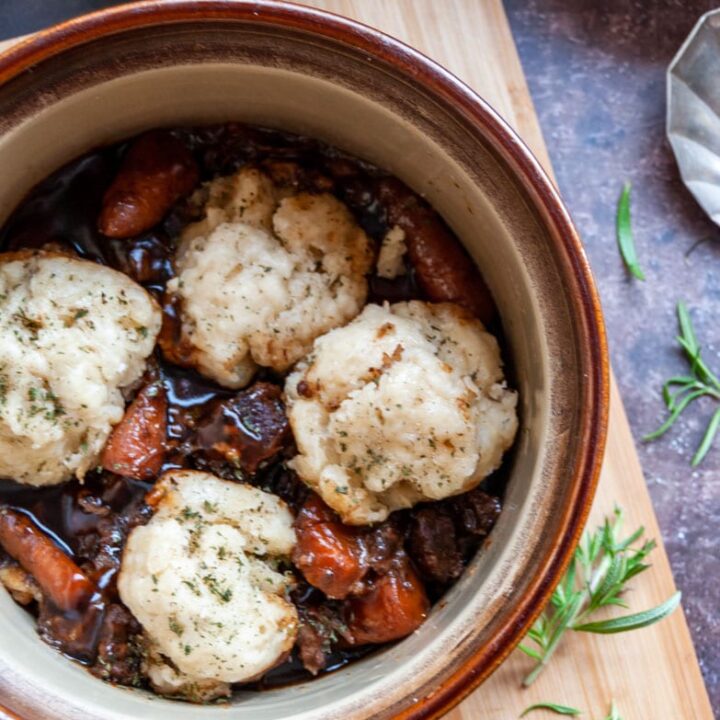 a brown ceramic bowl of beef stew with carrots and herb dumplings on a wooden board with fresh rosemary sprigs