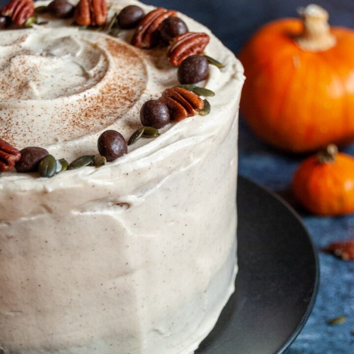 a close up picture of a three layer pumpkin cake covered in cream cheese frosting and topped with chocolate covered coffee beans, pecan nuts and pumpkin seeds.