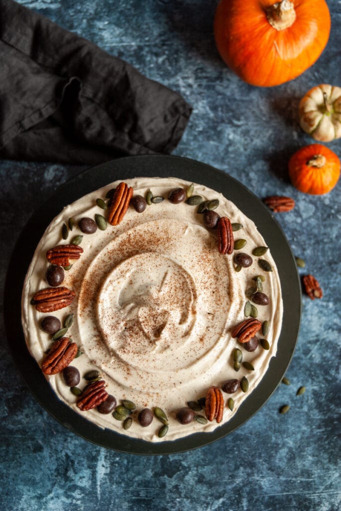 an overhead photo of a pumpkin layer cake covered in cream cheese frosting and decorated with pecan nuts, chocolate coffee beans and pumpkin seeds.