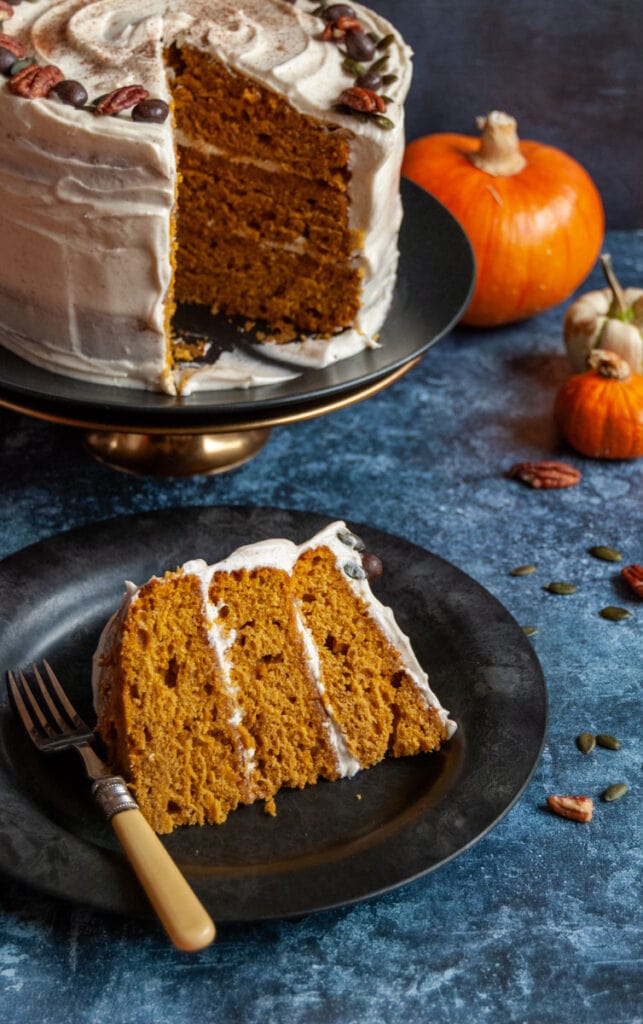 a slice of pumpkin cake covered in cream cheese frosting on a black plate.  A large pumpkin cake on a black plate and gold cake stand sits in the background.