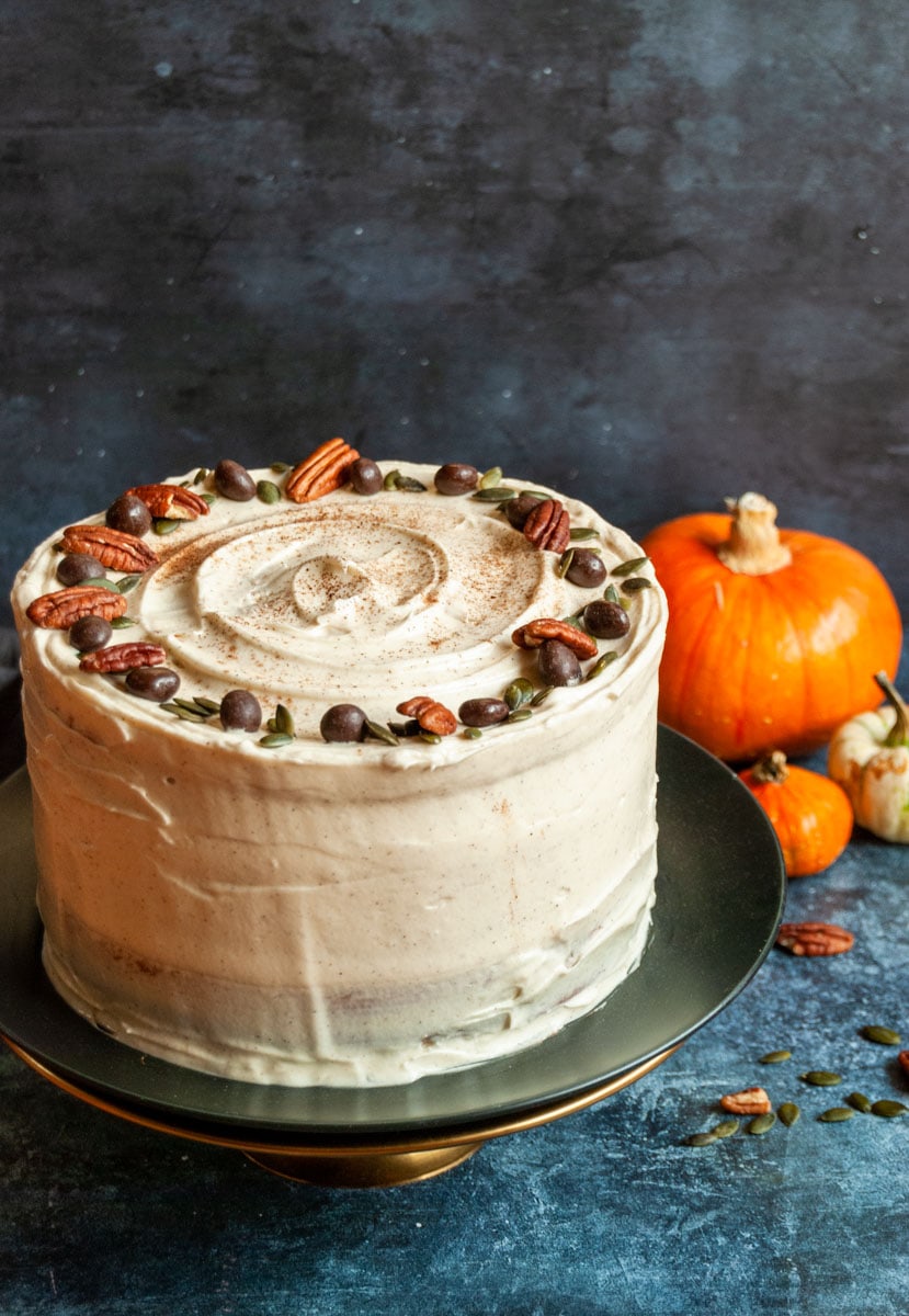 a pumpkin cake covered in cream cheese frosting and topped with pecan nuts, pumpkin seeds and chocolate covered coffee beans on a black plate and gold cake stand.