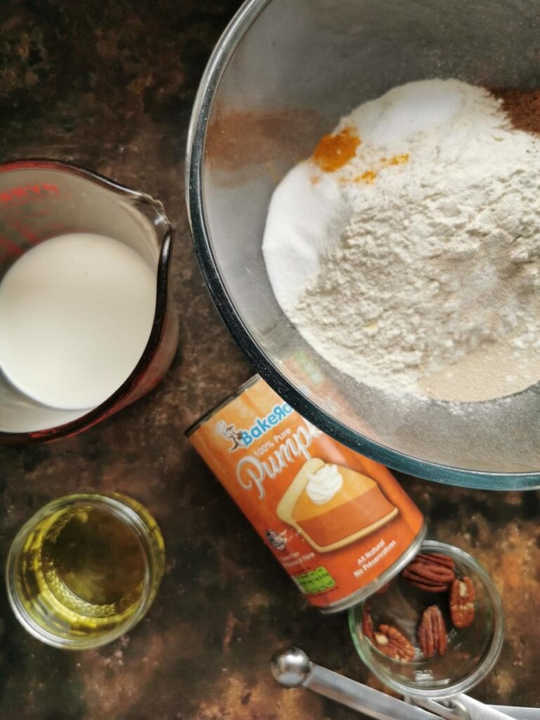 a bowl of flour, pumpkin pie spice, a tin of pumpkin puree, a jug of milk, a pot of pecan nuts and a pot of oil.