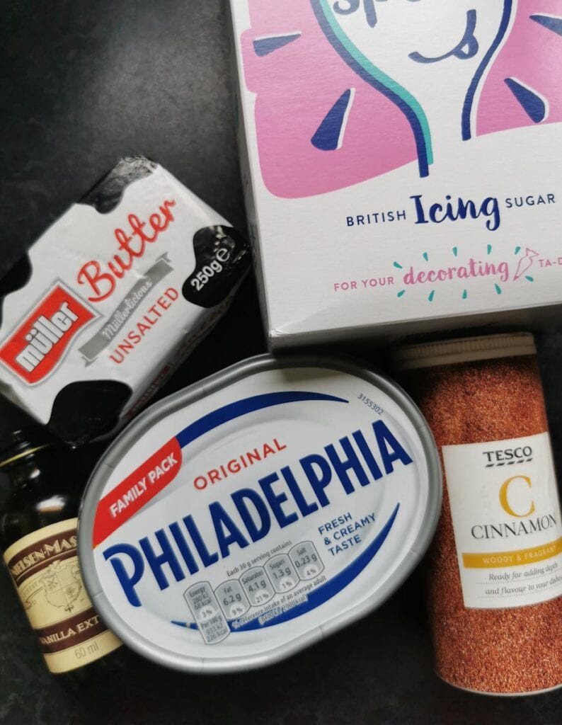 A box of icing sugar, a block of butter, a tub of cream cheese, vanilla extract and a tub of ground cinnamon.
