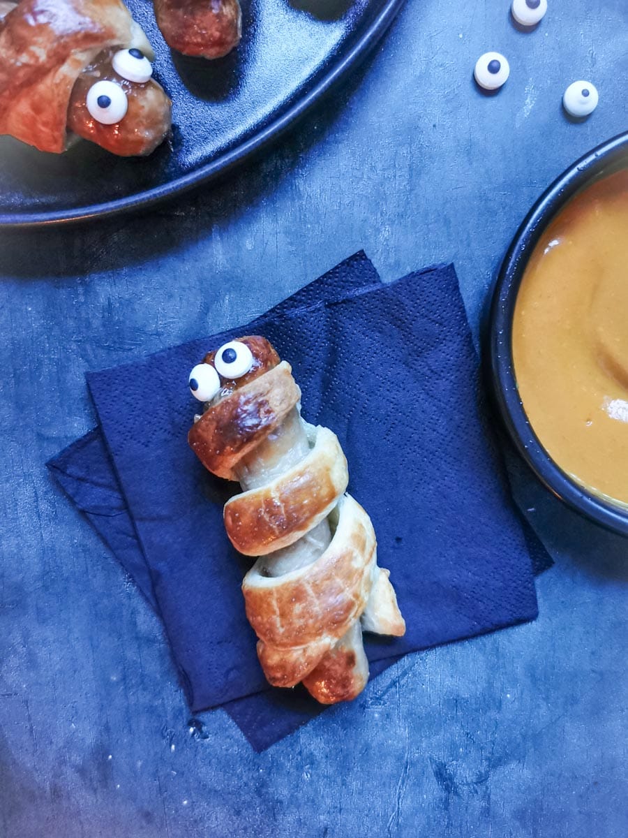 a sausage roll wrapped sausage with edible candy eyes to resemble Halloween "mummies" on a black napkin with a pot of spicy mustard dip.