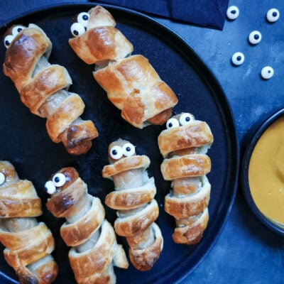 puff pastry wrapped sausages with edible eyes to resemble spooky "mummies" on a black plate, a black pot of yellow mustard dip and black napkins.