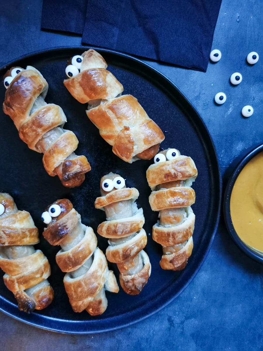 puff pastry wrapped sausages with edible eyes to resemble spooky "mummies" on a black plate, a black pot of yellow mustard dip and black napkins.