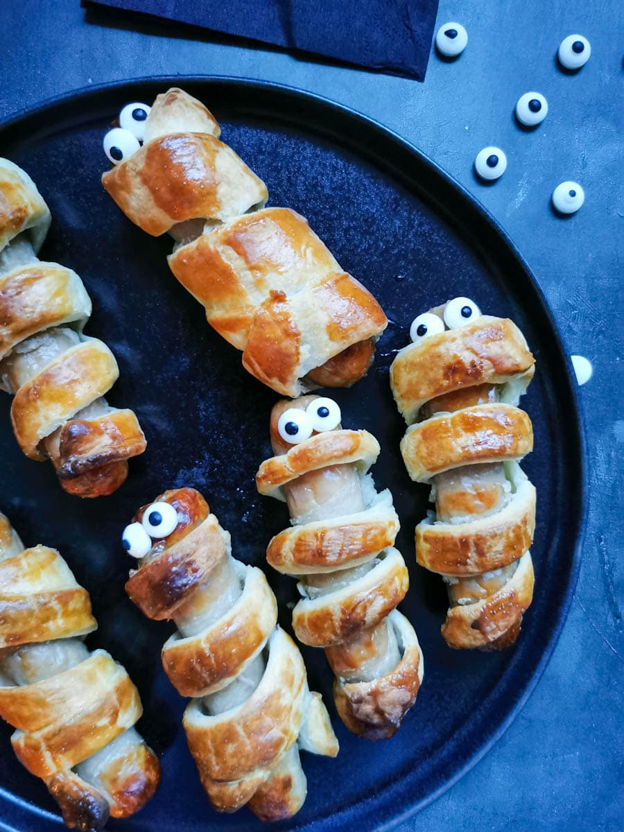 a black plate of puff pastry wrapped sausages with edible eyes to resemble "mummies".