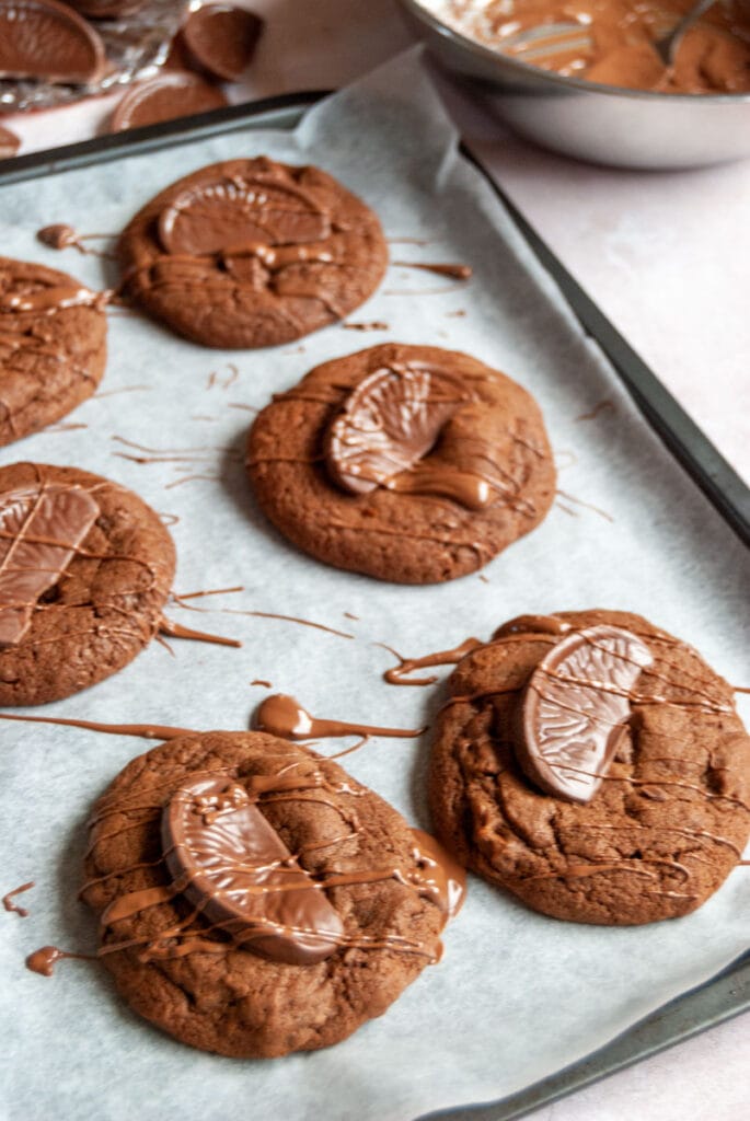 six chocolate cookies topped with a piece of chocolate orange and drizzled with melted milk chocolate on a baking tray.