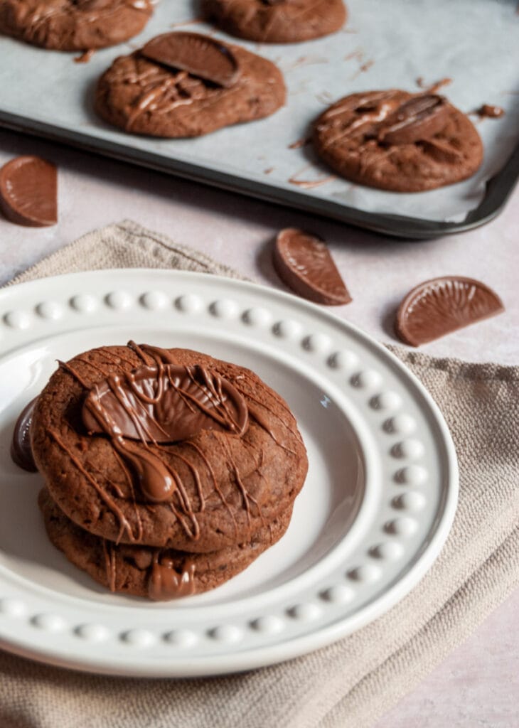 two chocolate orange cookies topped with a Terry's chocolate orange segment on a white plate.  More cookies can be seen on a baking tray in the background.