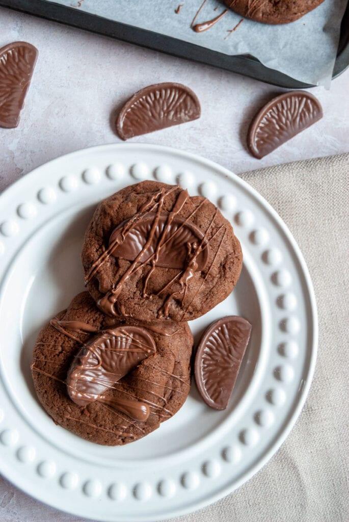 two chocolate cookies topped with a Terry's chocolate orange segment and drizzled with melted chocolate on a white plate. 