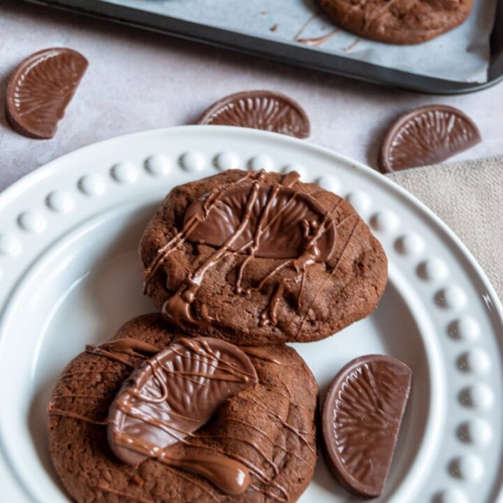 two large chocolate cookies topped with a Terry's chocolate orange piece and drizzled with melted chocolate on a white plate. More cookies can be partially seen on a baking tray in the background.