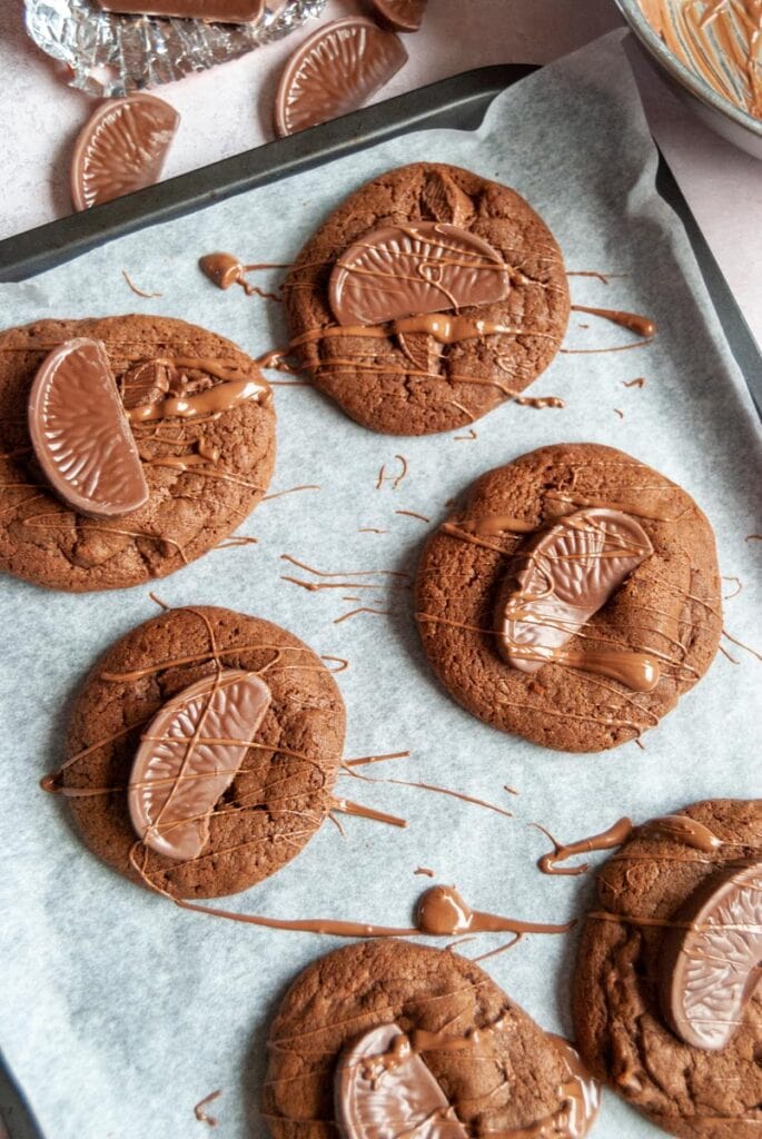 six chocolate cookies topped with a Terry's chocolate orange segement and drizzled with melted chocolte on a baking tray.