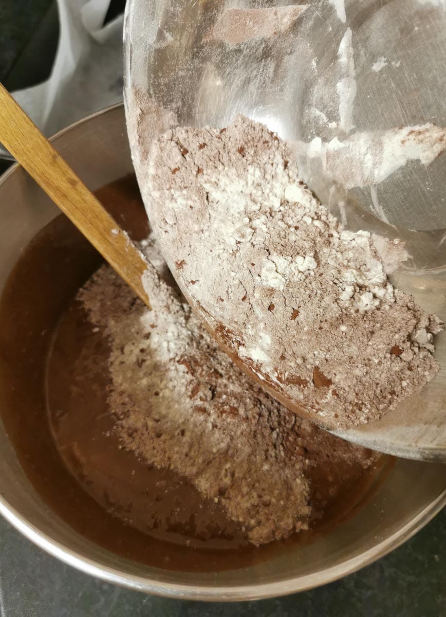 flour and cocoa powder being folded into a bowl of melted butter, eggs, sugar and chocolate.