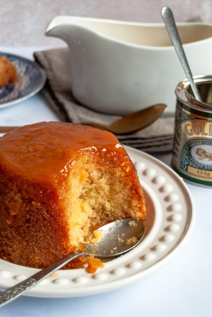a steamed sponge pudding with golden syrup and a silver spoon on a white plate.  A tin of golden syrup and a white jug of custard can be partially seen in the background.