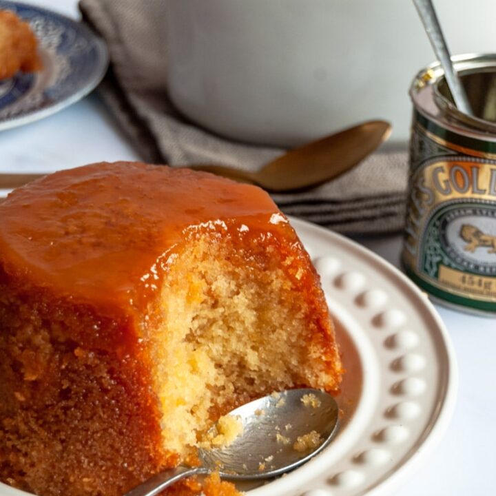 a steamed sponge pudding with golden syrup and a silver spoon on a white plate. A tin of golden syrup and a white jug of custard can be partially seen in the background.