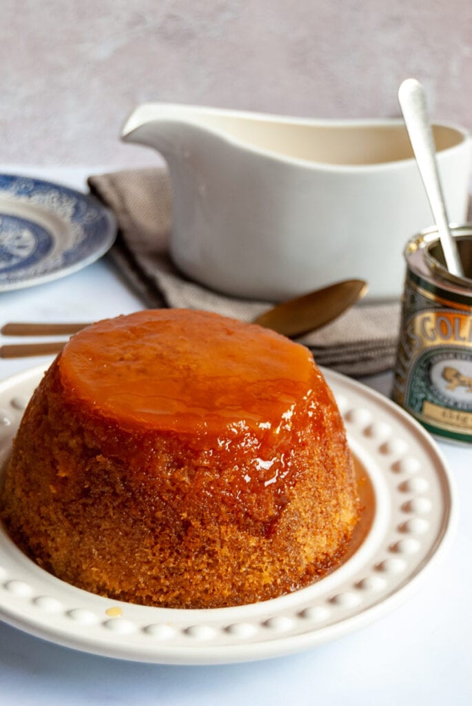 a steamed sponge pudding with golden syrup on a white serving plate, a tin of golden syrup, a white jug of custard and a blue willow pattern plate.