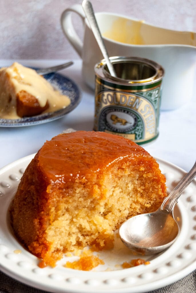 a steamed sponge pudding with golden syrup on a white plate with a large silver spoon.  A tin of golden syrup and a blue plate with a slice of sponge with custard can be seen in the background.