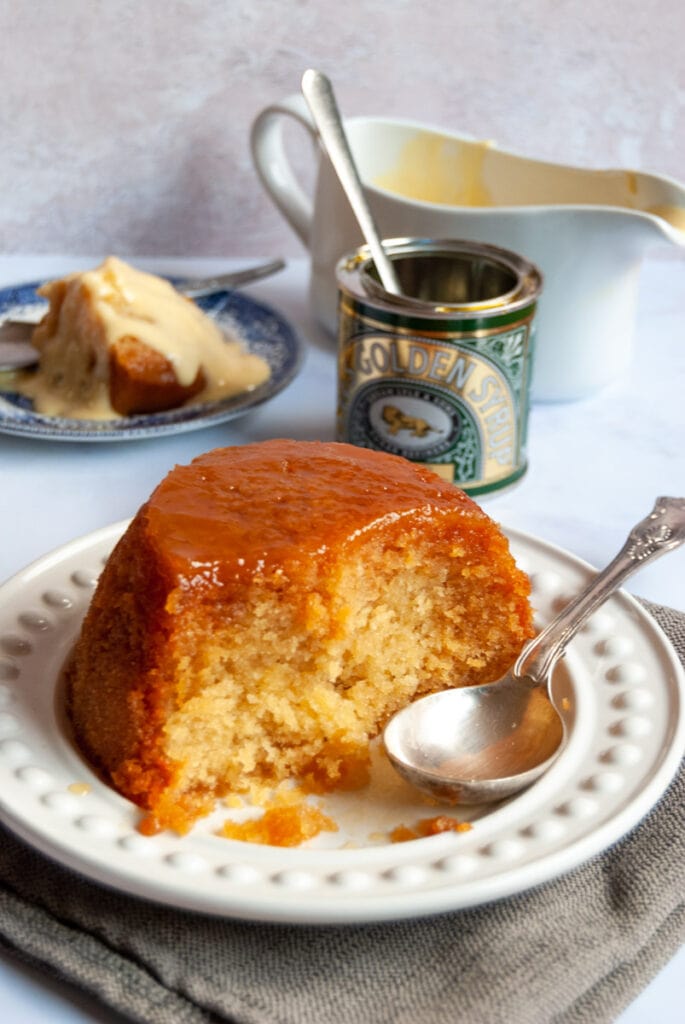 a sponge pudding with golden syrup on a white plate with a silver serving spoon, a tin of golden syrup and a white jug of custard.