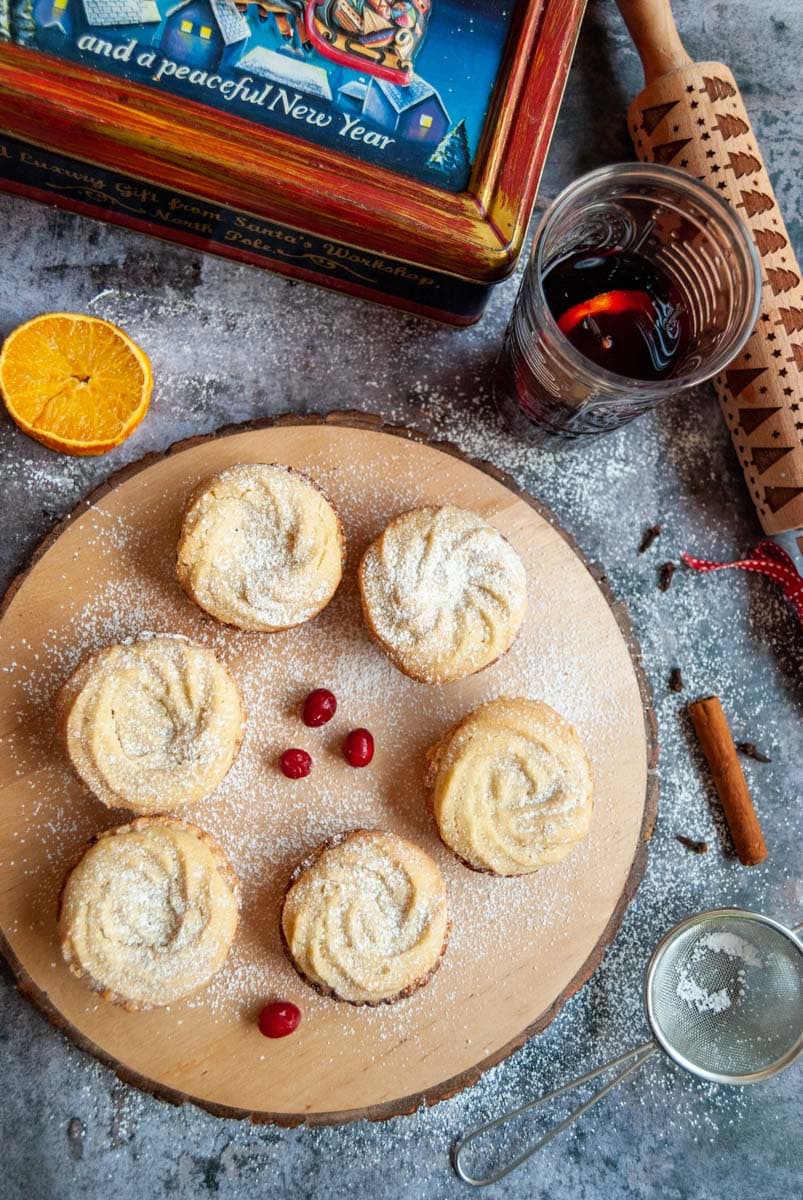 Six mince pies topped with a Viennese whirl on a wooden board dusted with icing sugar, a Christmas box with a picture of Santa on his sleigh, a glass of mulled wine and a rolling pin.