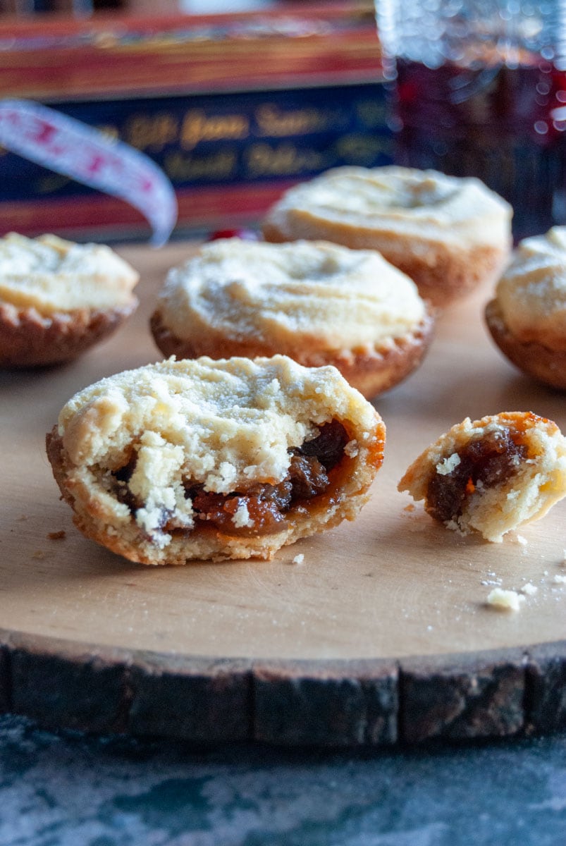 a close up picture of a mince pie with a crumble viennese whirl topping on a wooden serving board. More mince pies can be seen in the background.