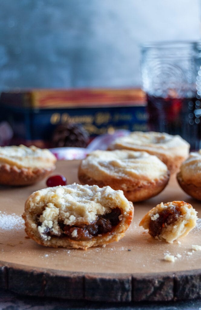 a mince pie broken in half to reveal the mincemeat filling on a wooden board. More mince pies can be seen in the background.