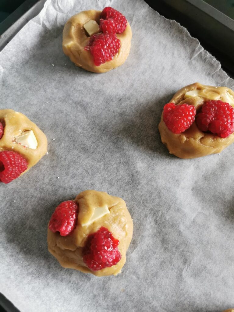 four unbaked cookies with white chocolate chunks and fresh raspberries on a lined baking sheet.