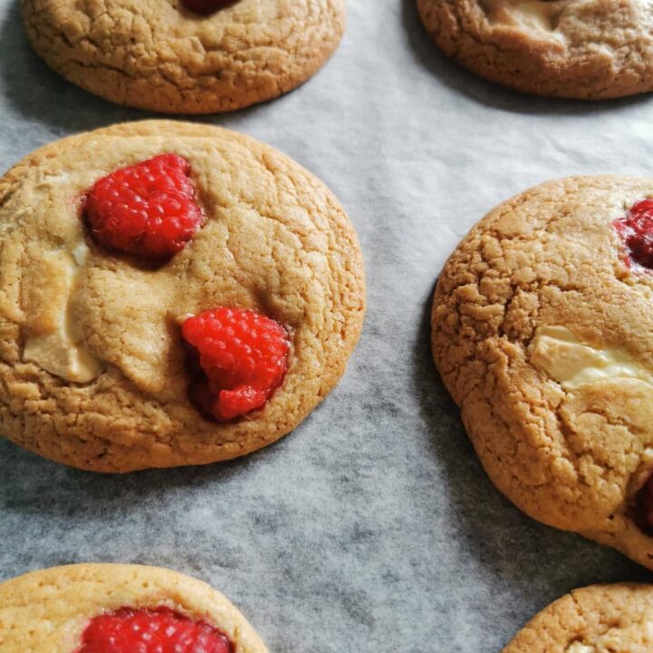 six large cookies with white chocolate chunks and fresh raspberries on a lined baking tray.