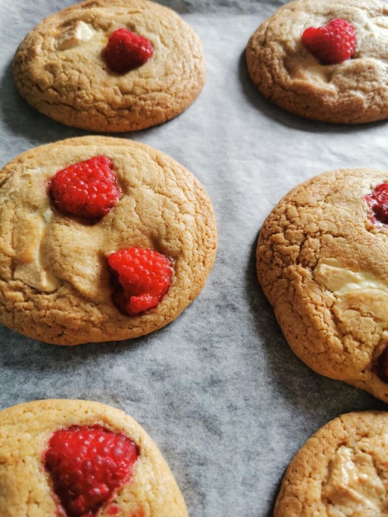 six large cookies with white chocolate chunks and fresh raspberries on a lined baking tray.