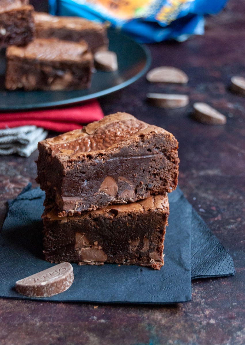 two chocolate brownies with milk chocolate chunks on a black paper napkin. A plate of brownies can be partially seen in the background.