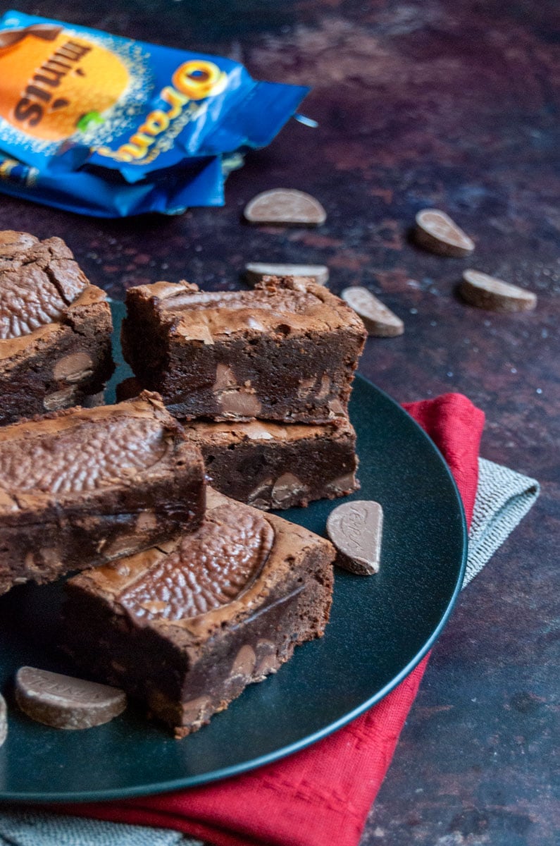 a black plate of chocolate brownies with milk chocolate orange chunks and a packet of chocolate orange sweets.