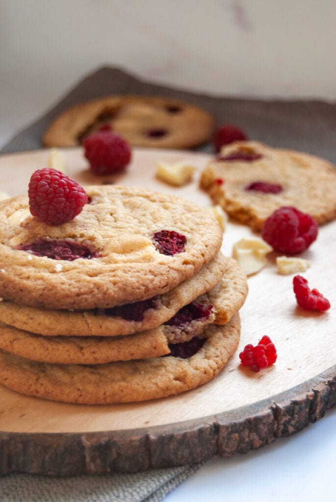 a stack of white chocolate cookies with fresh raspberries on a wooden serving board.  More cookies can be seen in the background.