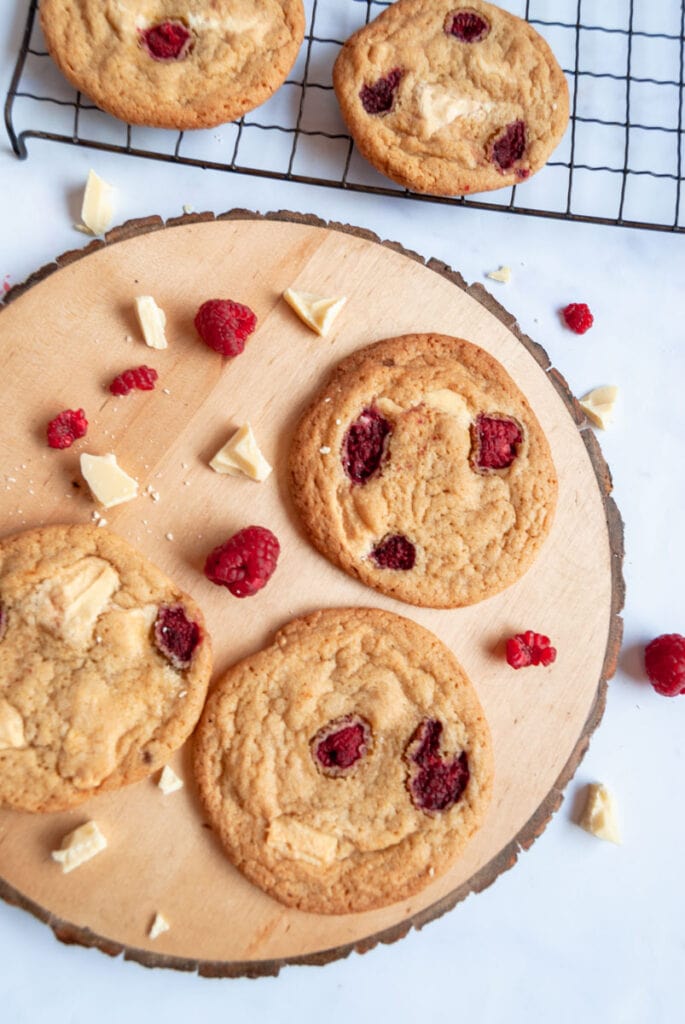 three white chocolate cookies with raspberries on a wooden board with fresh raspberries and white chooolate chunks.