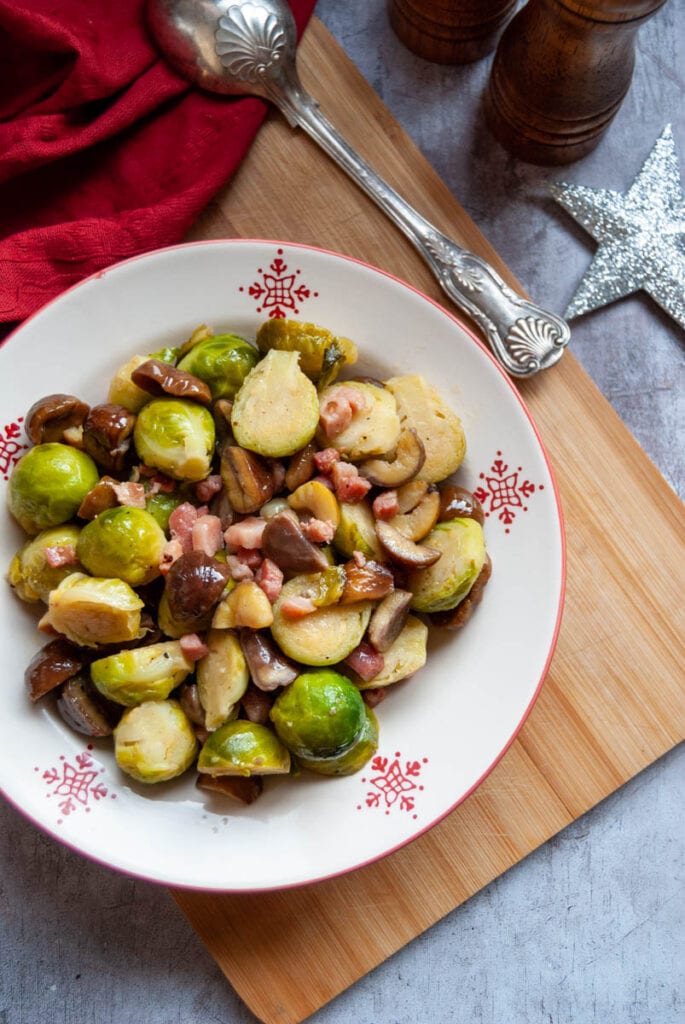 a white and red bowl of brussels sprouts with bacon and chestnuts on a wooden board.