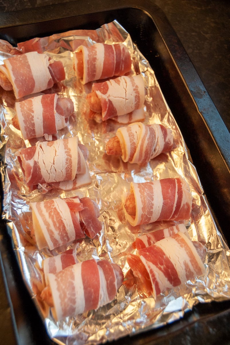 ten small pork sausages wrapped in bacon on a tin foil lined baking tray