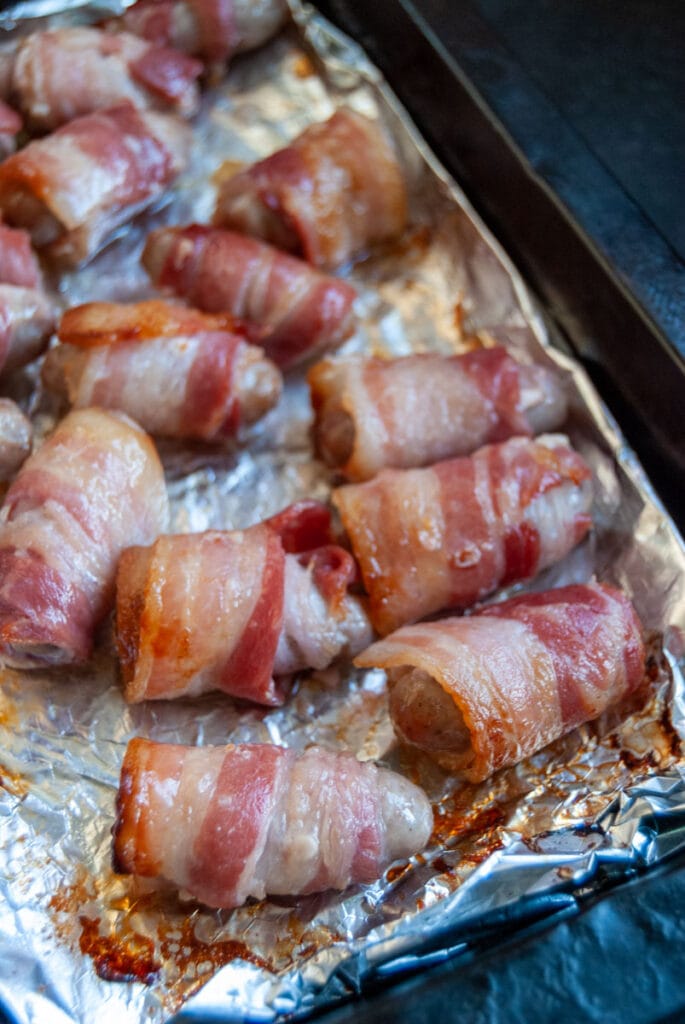 cooked sausages wrapped in bacon on a foil lined baking tray.