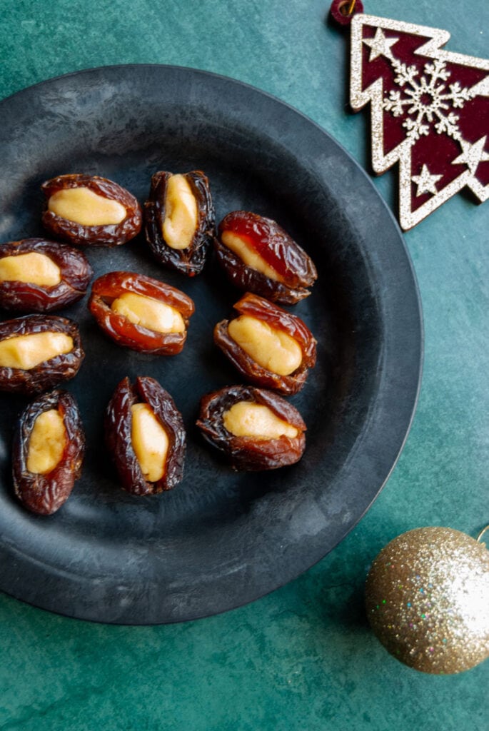 a black plate of marzipan stuffed medjool dates on a dark green background with red and gold Christmas decorations.