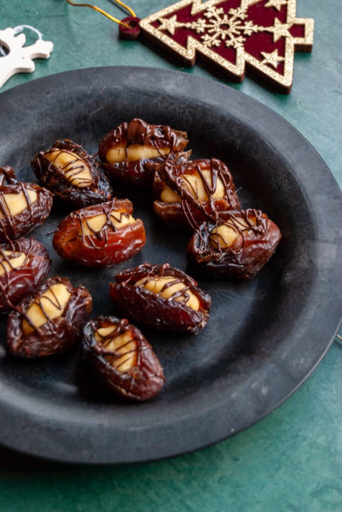 a black plate of marzipan stuffed dates drizzled with dark chocolate on a dark green backdrop.