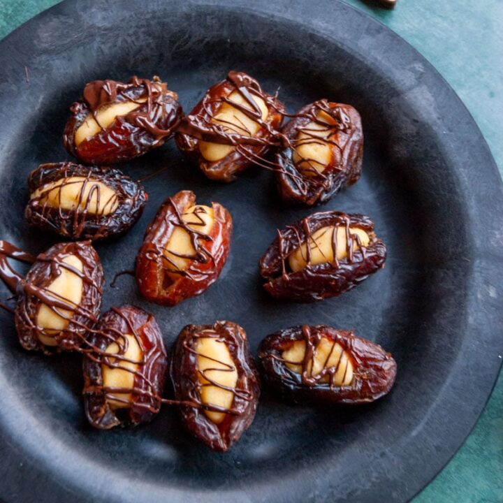 a black plate of medjool dates stuffed with marzipan and drizzled with dark chocolate on a dark green background with Christmas decorations.