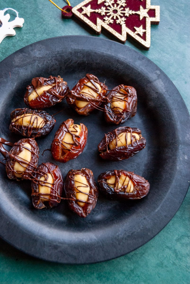 a black plate of medjool dates stuffed with marzipan and drizzled with dark chocolate on a dark green background with Christmas decorations.