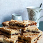 shortbread squares filled with mincemeat and topped with flaked almonds on a wooden board with a wooden star Christmas decoration. A small white milk jug and a white and silver tea cup with a spoon sits behind the shortbread bars.