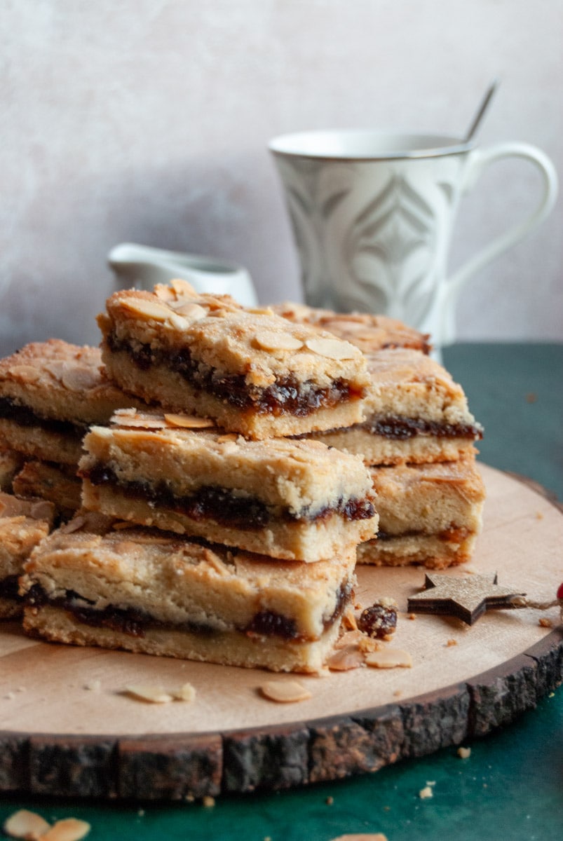 shortbread bars filled with mincemeat and topped with flaked almonds on a wooden board and a white and silver tea cup and a small white milk jug in the background