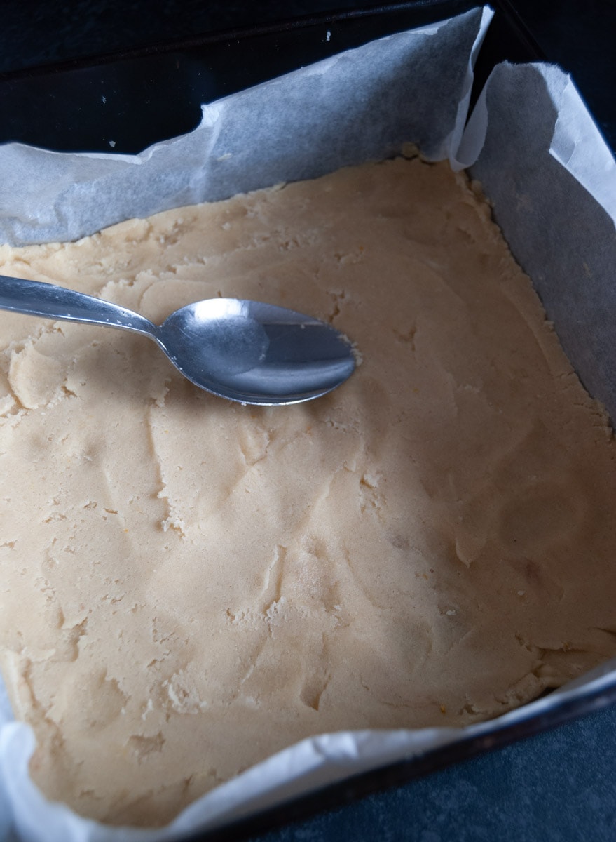 Shortbread dough in a square lined baking tin with a silver spoon.