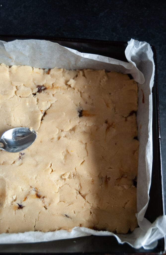 unbaked shortbread dough with a mincemeat filling being pressed into a square baking tin with a silver spoon.
