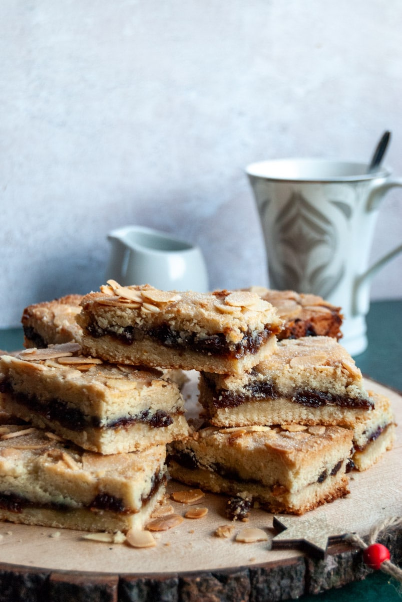 shortbread squares filled with mincemeat and topped with flaked almonds on a wooden board with a wooden star Christmas decoration. A small white milk jug and a white and silver tea cup with a spoon sits behind the shortbread bars.