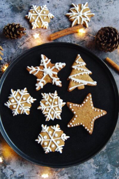 a black plate of gingerbread cookies cut into Christmas shapes and decorated with white icing. Pine cones, cinnamon sticks and fairy lights surround the plate.