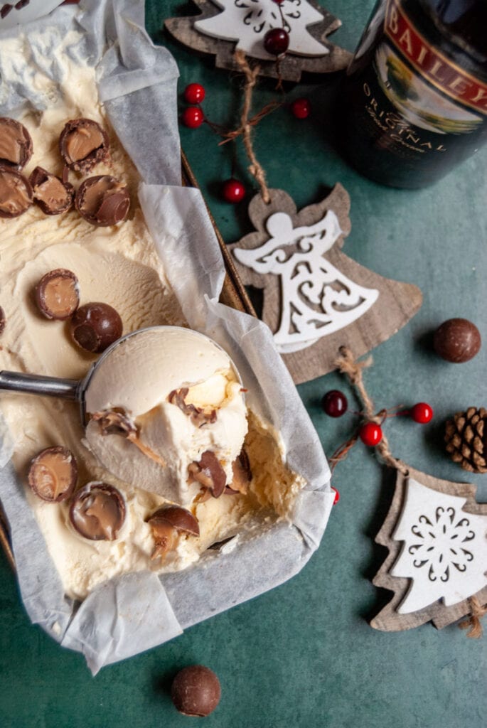 a loaf pan of ice cream topped with Baileys chocolate truffles, A wooden white and red Christmas decoration and a bottle of Baileys Irish cream liqueur.