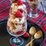 Four dessert glasses filled with whipped double cream, raspberries and toasted oats and nuts on a black plate with two gold serving spoons.