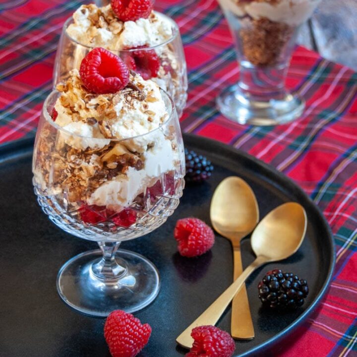 two dessert glasses filled with whipped cream,fresh raspberries and toasted oats on a black plate with two gold spoons and fresh raspberries and a tartan tablecloth.